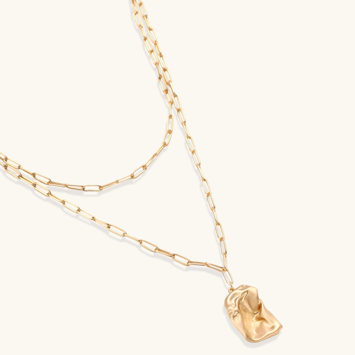 Layered gold paperclip chain necklace with organic sculptural pendant in warm lighting on cream background