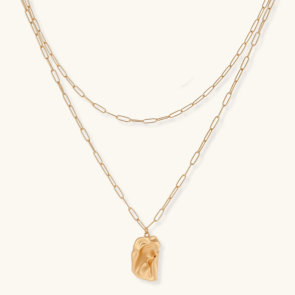 Layered gold paperclip chain necklace with organic sculptural pendant in warm lighting on cream background
