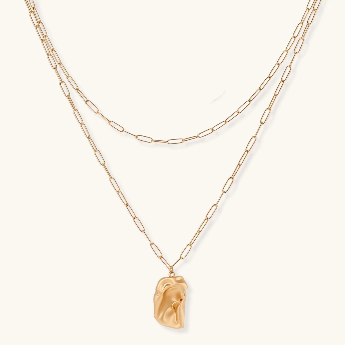 Layered gold paperclip chain necklace with organic sculptural pendant in warm lighting on cream background