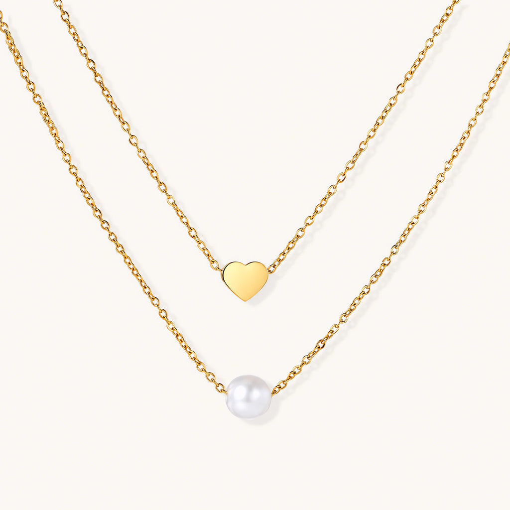 Two delicate gold-plated chains with heart charm and white pearl bead on cream background, minimal layered design.