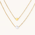 Two delicate gold-plated chains with heart charm and white pearl bead on cream background, minimal layered design.