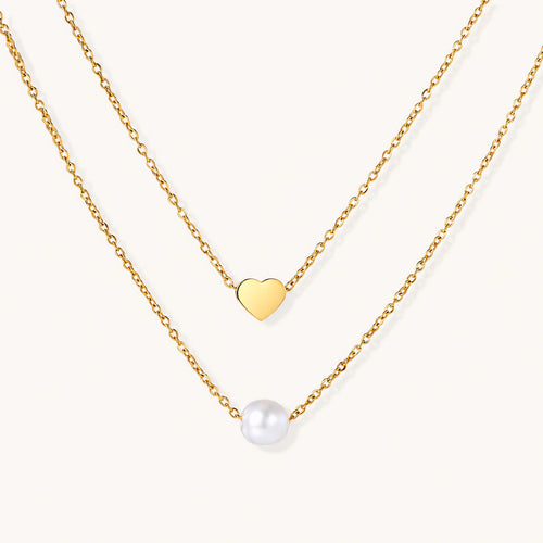 Two delicate gold-plated chains with heart charm and white pearl bead on cream background, minimal layered design.